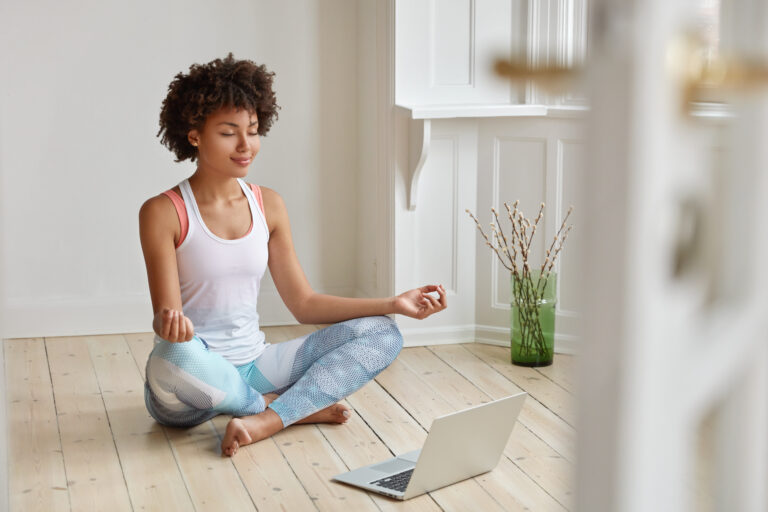 Calm lady with Afro hairstyle, dressed in sportswear, meditates on floor in empty room, listens spiritual practices lessons on laptop computer, poses in lotus pose, tries to relax